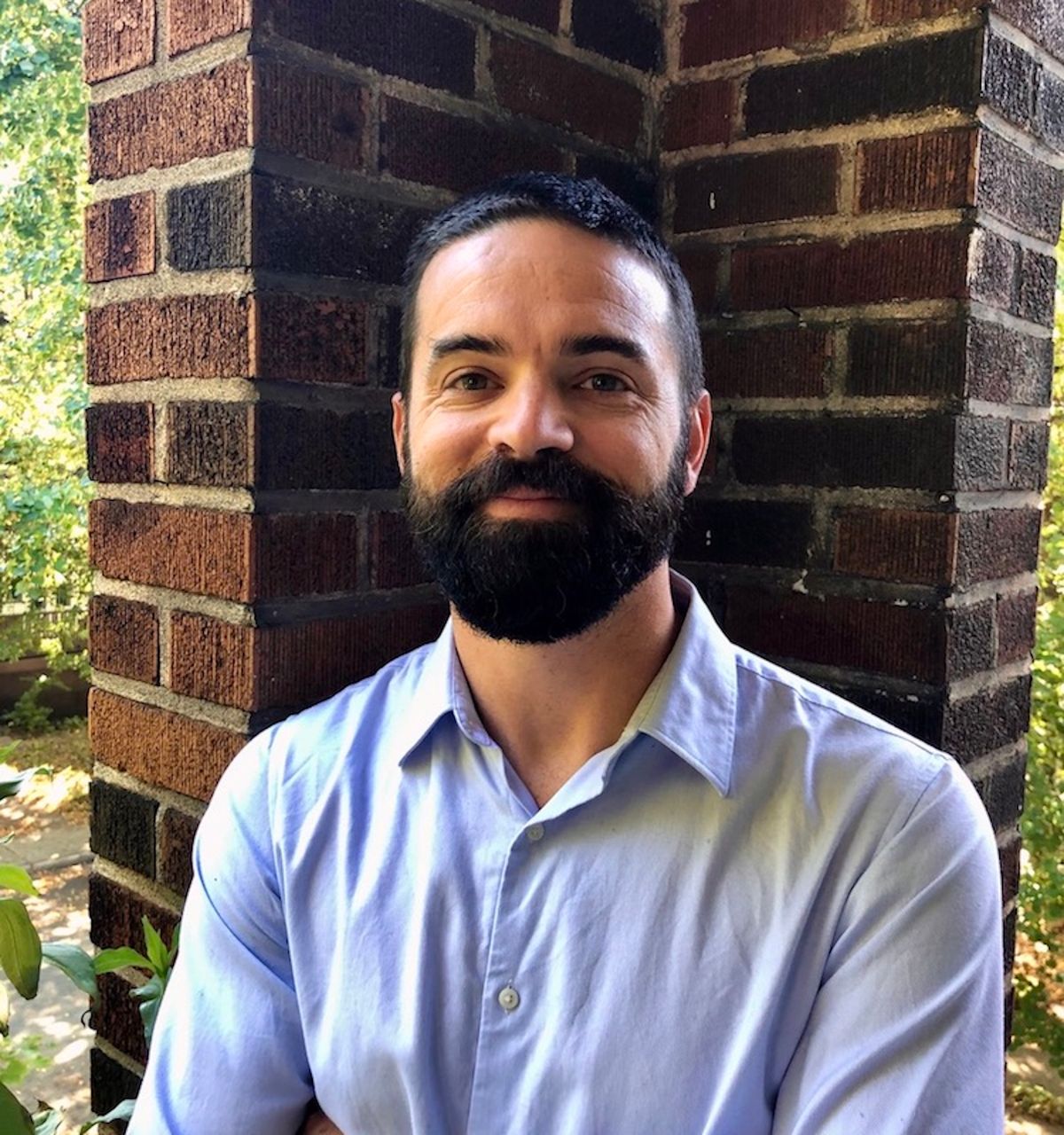 Matthew Harrington wears a blue collared shirt and stands outside in front of a brick wall. Matthew Harrington wears a blue collared shirt and stands outside in front of a brick wall.