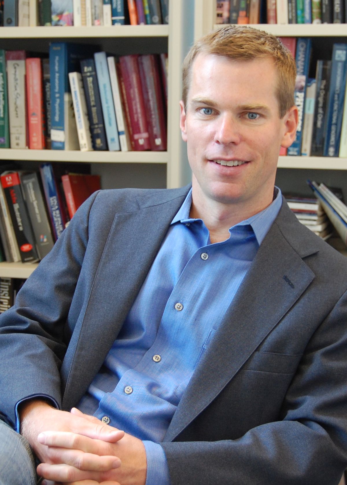 A man wearing grey suit sits in front of a bookshelf. A man wearing grey suit sits in front of a bookshelf.
