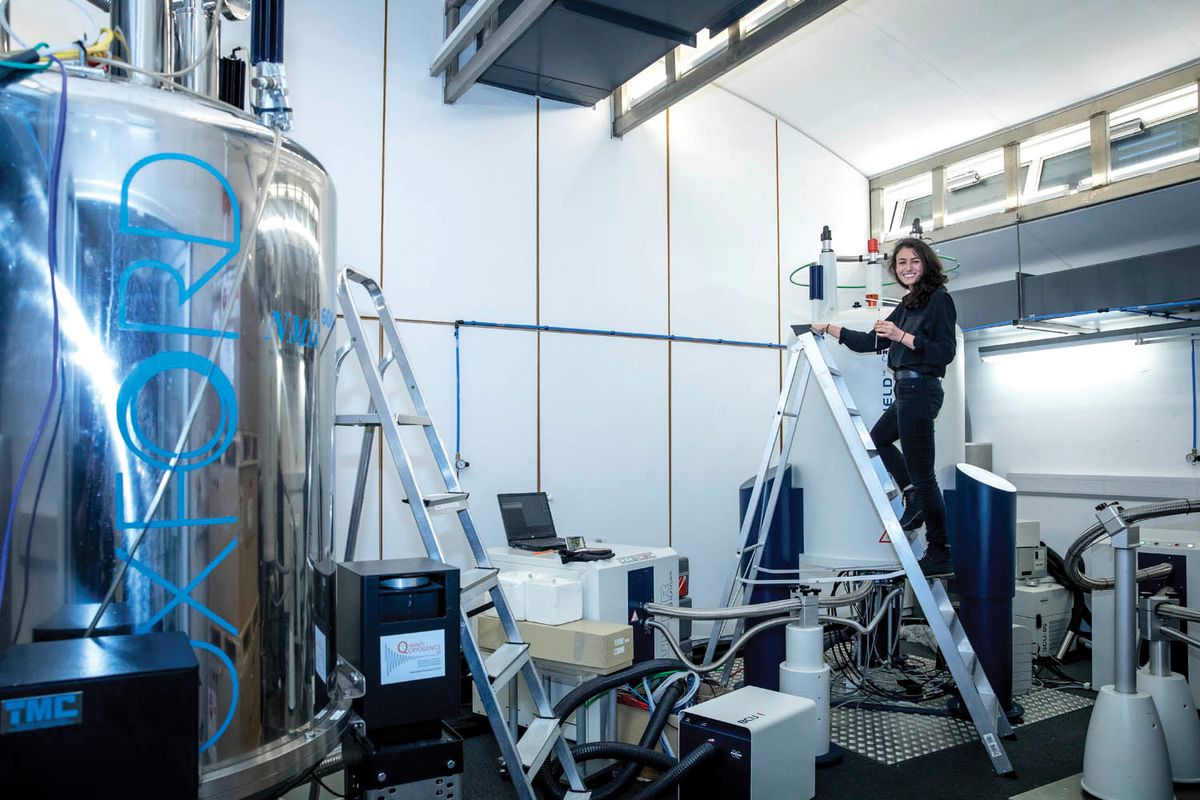 Photo of Gabriella Heller standing on a ladder next to a nuclear magnetic resonance spectrometer. Photo of Gabriella Heller standing on a ladder next to a nuclear magnetic resonance spectrometer.
